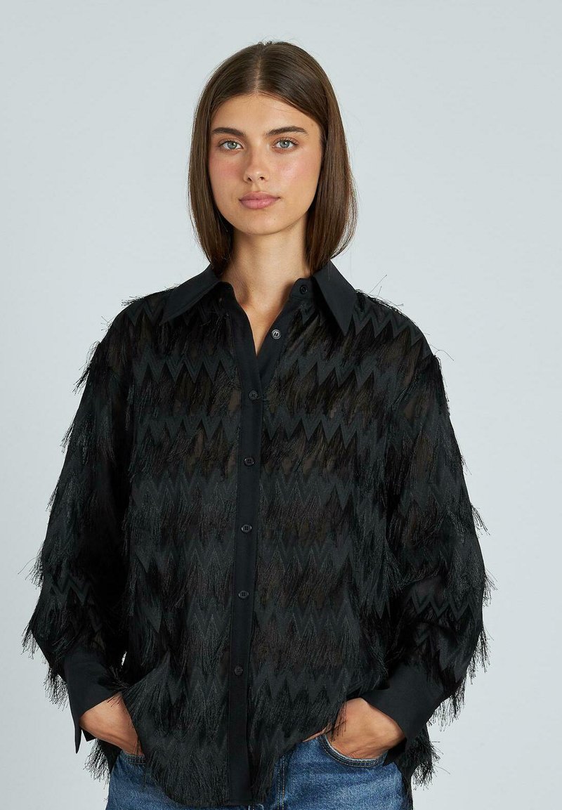 Camisa negra con un patrón en zigzag, fabricada en un material ligero con detalles de flecos, y un cuello con cierres de botón.