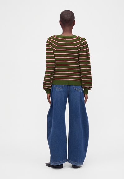 Personne debout de dos, portant un pull rayé vert et rouge avec des manches bouffantes et un jean bleu à jambes larges.