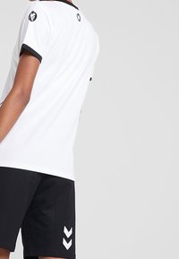 Bílá košile s krátkými rukávy s černými detaily a logem na rukávu, doplněná černými šortkami s bílým chevronovým vzorem.