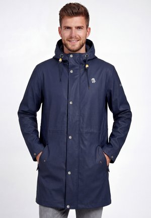 Mężczyzna z jasnym zarostem, ubrany w granatową przeciwdeszczową kurtkę z kapturem, z rękami w kieszeniach, uśmiechający się, stojący na białym tle.