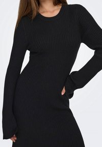 Femme portant une robe noire côtelée à manches longues, taille cintrée et encolure ronde, posant avec une main sur la hanche.
