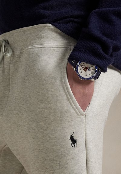 Gaiši pelēkas sporta bikses no mīksta auduma, ar v formas jostasvietu un mazu melnu Polo logotipu. Uz cilvēka ir tumši zils džemperis un pulkstenis.