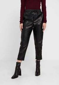 Zwarte faux leren broek met een elastische tailleband en trekkoord, met een cropped pasvorm die net boven de enkel eindigt, gedragen met zwarte haklaarzen.