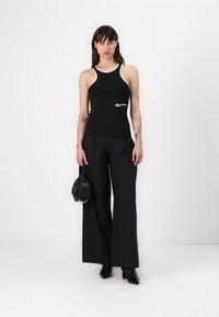 Top de tirantes negro ajustado con cuello y logotipo blanco, combinado con pantalones anchos negros. El conjunto incluye un bolso estructurado negro y botines.