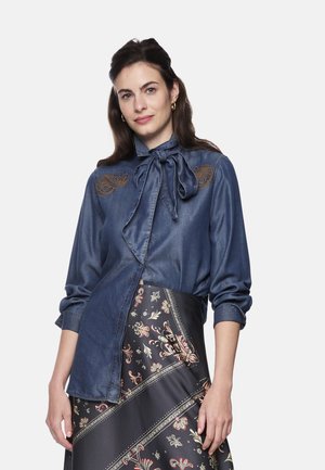 Denim blouse in deep blue, featuring a bow tie collar and intricate embroidery on the shoulders, paired with a patterned black skirt.