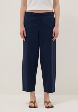 Femme portant un pantalon large court bleu marine avec une taille à cordon et des sandales marron à brides, debout devant un fond uni.