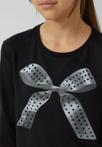 Schwarzes T-Shirt mit großem silbernem Schleifenmotiv, das perforierte Details und schwarze Akzente aufweist, Rundhalsausschnitt, lange Ärmel.