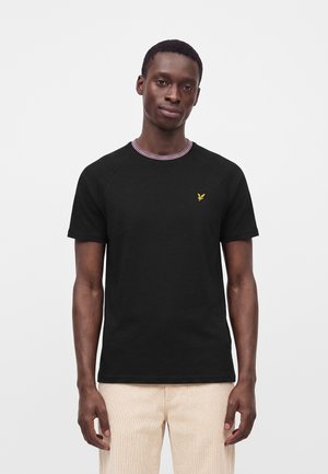 Mann trägt ein schwarzes kurzärmeliges Hemd mit kleinem gelbem Vogel-Logo auf der Brust und hellbeige Hose, steht vor weißem Hintergrund.