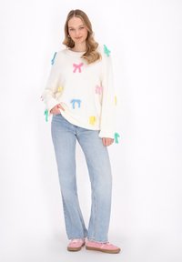 Weißer Strickpullover verziert mit bunten Filzschleifen in Rosa, Blau, Grün und Gelb, kombiniert mit hellblauen Schlagjeans und rosa Schuhen.