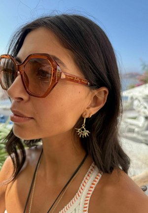 Frau mit dunklen Haaren, die große achteckige braune Sonnenbrillen, sonnenförmige Ohrringe mit bunten Steinen und ein weißes Stricktop im Freien trägt.