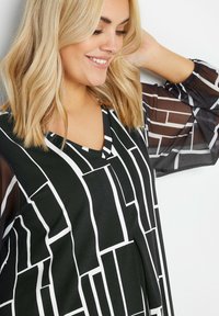 Top con motivo bianco e nero caratterizzato da una scollatura a V e maniche a palloncino trasparenti. Il design è composto da forme geometriche con linee a contrasto.