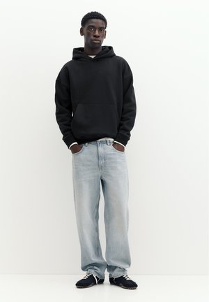 Junger Mann steht mit Händen in den Taschen, trägt schwarzen Hoodie, hellblaue weite Jeans und schwarze Sneakers vor einem schlichten weißen Hintergrund.