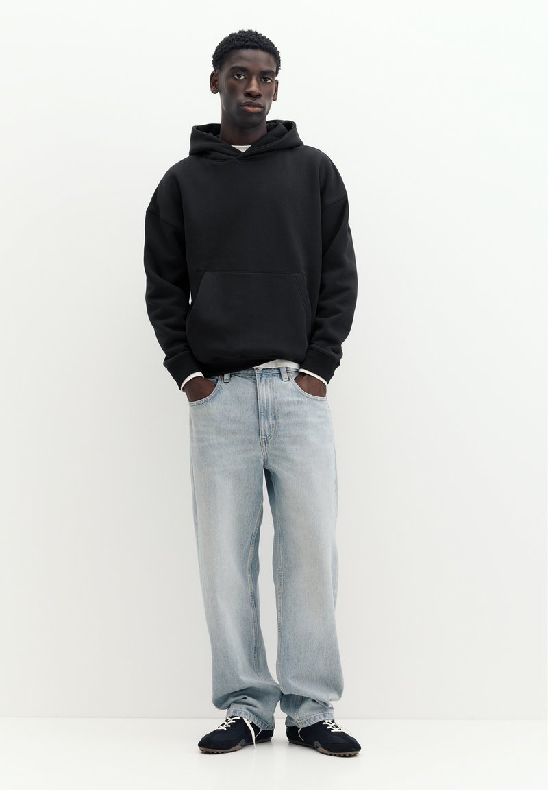 Jeune homme debout les mains dans les poches, portant un sweat à capuche noir, un jean large bleu clair et des baskets noires sur fond blanc uni.