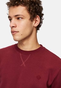 Burgundi-sweatshirt, jossa on pyöreä kaula-aukko, teksturoitu ommeltyö ja pieni logo rinnassa, valmistettu pehmeästä ja sileästä kankaasta.
