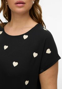 Vrouw draagt een zwart shirt met witte hartjes, korte mouwen en een ronde hals, waarbij nek, kin en een deel van het haar zichtbaar zijn.