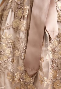 Robe beige avec une broderie florale dorée complexe, des accents en perles et un ruban en satin. Tissu texturé avec des éléments superposés et des détails délicats.
