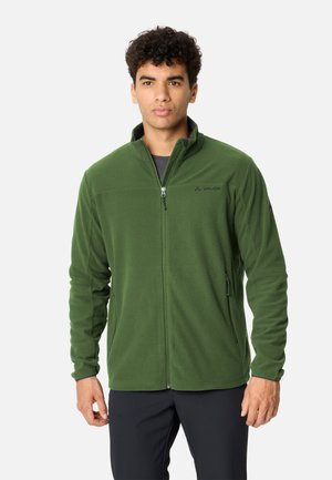Junger Mann, der eine grüne Fleecejacke mit Reißverschluss und schwarze Hosen trägt, vor einem einfachen hellen Hintergrund stehend.