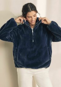 Pulover din fleece bleumarin cu guler înalt, fermoar frontala și buzunare laterale; are o textură moale și o croială lejeră.