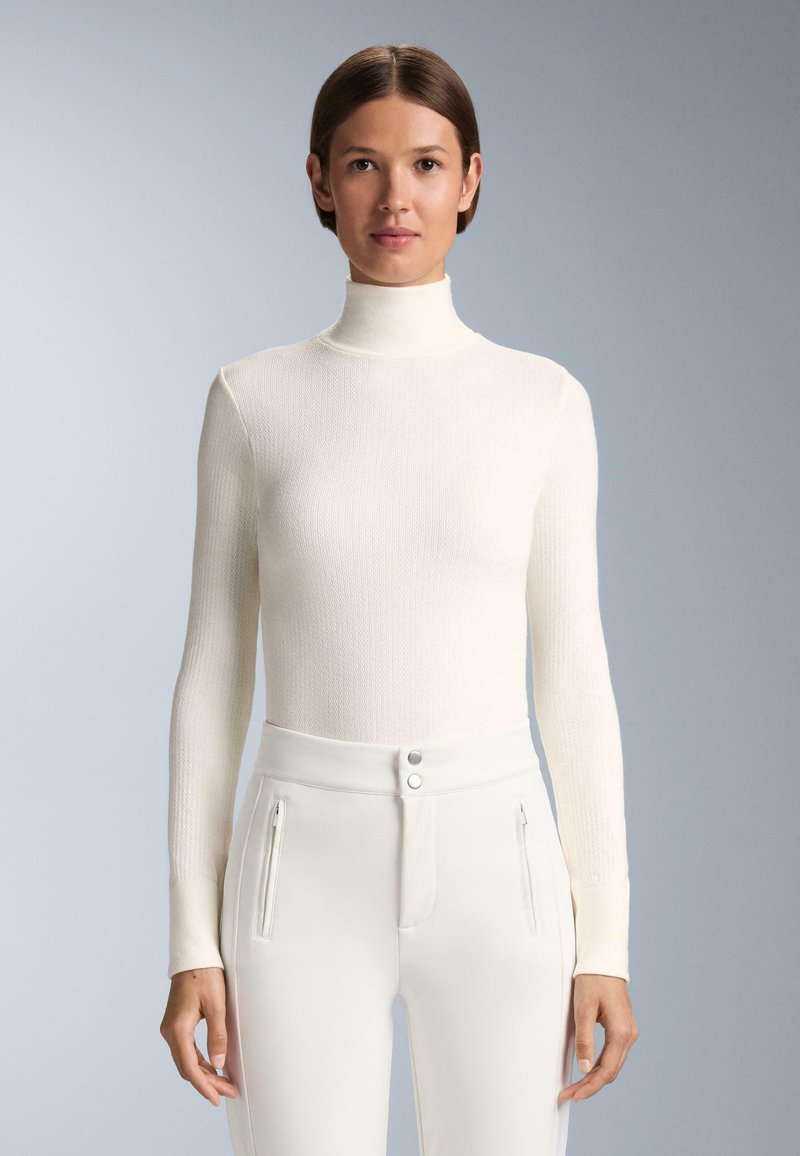 Witte geribde turtleneck trui met lange mouwen, gecombineerd met hoogziende witte broek met ritsen en knoopsluitingen.