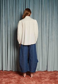 Žena s dlhými vlasmi stojí čelom k svetlomodrým závesom, oblečená v voľnej bielej blúzke, širokých námorníckych nohaviciach s malými bielymi bodkami a bielych podpätkoch.