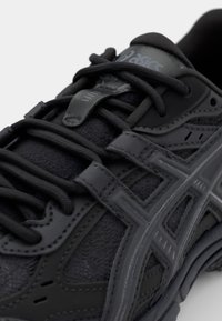 Svart sportskor med texturerad läder- och meshöverdel, med ett framträdande snörningssystem och en grå logotyp på tungan.