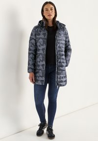 Gewatteerde jas in donkergrijs met een subtiel ton-sur-ton patroon, voorzien van een capuchon, ritssluiting en elastische manchetten. Draagt over een zwart shirt en jeans.