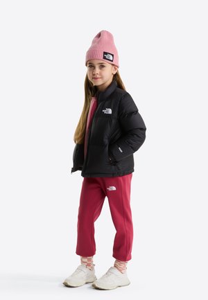 Jeune fille aux longs cheveux portant un bonnet en laine rose, une doudoune noire, un pantalon rose, des chaussettes à motifs et des baskets blanches, debout les mains dans les poches.