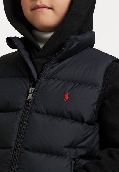 Colete puffer azul-marinho com colarinho alto, fecho de correr e logótipo bordado em vermelho. Usado sobre um hoodie preto. Textura lisa e acolchoada.