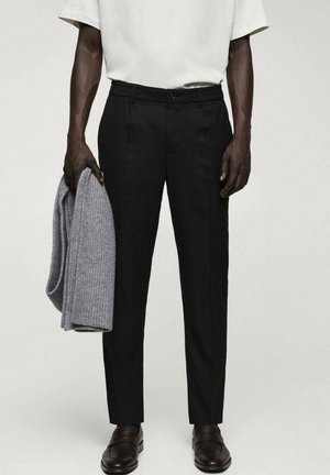 Mann trägt schwarze, maßgeschneiderte Hose, weißes Hemd, schwarze Slipper und hält einen gefalteten, grauen Strickpullover in der linken Hand vor einfachem Hintergrund.