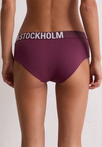 Burgunderfarbene Baumwoll-Hipster-Unterwäsche mit breitem elastischen Bund, der "STOCKHOLM" in hellgrau zeigt. Glatte Textur, figurbetontes Design.