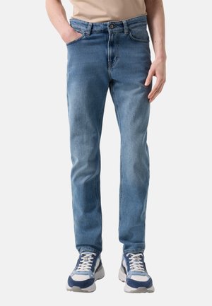 Mann, der hellblaue Jeans mit geradem Bein und blaue, weiße und graue Sneakers trägt, steht mit einer Hand in der Tasche vor weißem Hintergrund.