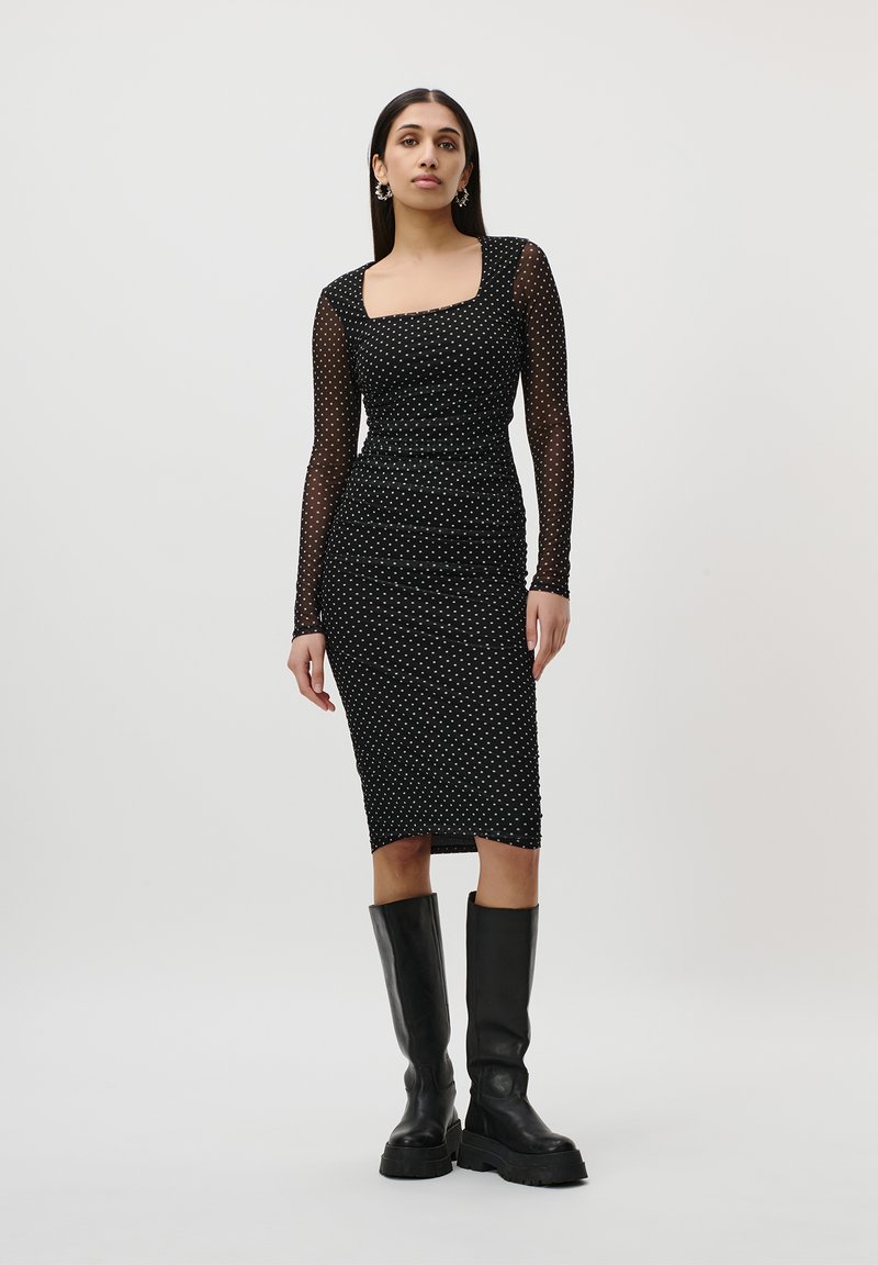 Abito nero a pois con scollatura quadrata, lunghe maniche trasparenti, design aderente e orlo al ginocchio, abbinato a stivali neri alti.