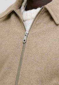 Chaqueta de mezcla de lana beige con cremallera en la parte delantera, cuello puntiagudo y herrajes de cremallera en plata. La textura parece suave y cálida.