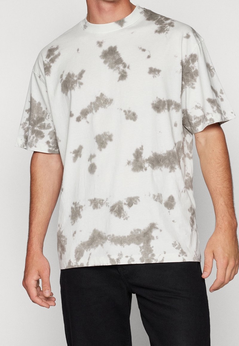 Homme portant un t-shirt blanc à manches courtes avec des motifs tie-dye gris et un pantalon noir sur un fond uni.