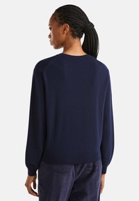 Femme avec des cheveux tressés en queue de cheval, portant un pull bleu marine et un pantalon noir côtelé, de dos contre un fond blanc.