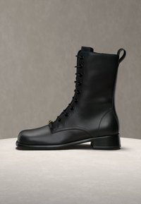 Bottine noire en cuir à lacets, talon bas, couture latérale et languette à l'arrière sur une surface claire.