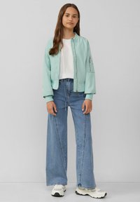 Mintgrüne Bomberjacke mit Reißverschluss, gerippten Bündchen und einer Seitentasche, kombiniert mit hellblauen, weit geschnittenen Jeans und weißen Sneakers.