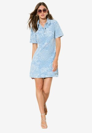 Kvinde iført lyseblå kortærmet blondekjole med blomster, krave, solbriller, guldøreringe og hvide højhælede sandaler med stropper.