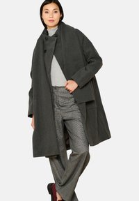 Cappotto di lana grigio scuro con una vestibilità ampia, collo grande e chiusura a doppi bottoni, abbinato a pantaloni lucidi in argento testurizzati e scarpe bordeaux.