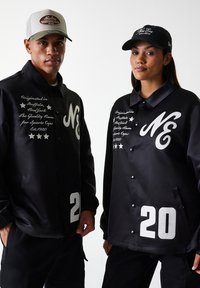 Black satin jackets feature white lettering and numbers. Jackets have button closures, two side pockets, and an embroidered logo on the chest.