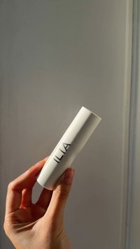 Prodotto cilindrico bianco con superficie liscia, etichettato "ILIA" con lettere scure. Tenuto in una mano su uno sfondo neutro.