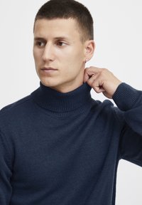 Sötétkék garbó pulóver bordás kötésű anyagból. Magas nyakkal és hosszú ujjakra készült, amely finom textúrát és kivágást mutat.