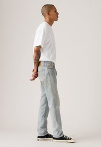 Άσπρο μπλουζάκι με λαιμόκοψη τύπου crew, ανοιχτό μπλε τζιν παντελόνι και μαύρα πάνινα αθλητικά παπούτσια. Ξεβαμμένο τζιν με ίσια γραμμή και ελάχιστες λεπτομέρειες.