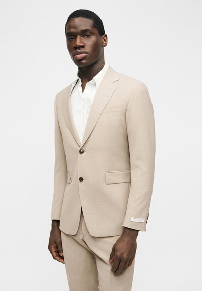 Μαύρος άνδρας φοράει μπεζ κοστούμι με καφέ κουμπιά πάνω από λευκό πουκάμισο, στέκεται με ουδέτερη έκφραση μπροστά σε απλό φωτεινό φόντο.