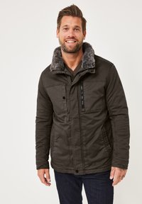 Dunkelgrüne Jacke mit einem grauen Kunstfellkragen, ausgestattet mit Reißverschluss- und Knopfentaschen sowie einem strukturierten Design. Über einem braunen Pullover getragen.