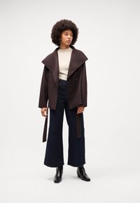 Cappotto in misto lana marrone con colletto ampio, chiusura a bottoni e cintura, abbinato a un top a coste color crema e jeans larghi scuri. Stivali neri.