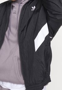 Svart vindjacka med vit sidopanel, lättviktsmaterial, har en broderad logotyp på bröstet, elastiska ärmslut och en dragkedja fram.