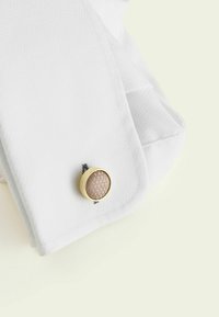 Weißer Stoff mit Struktur und einem goldfarbenen runden Knopf, der eine erhabene, weich beige Mitte aufweist. Schwarze Stickdetails in der Nähe des Knopfs.