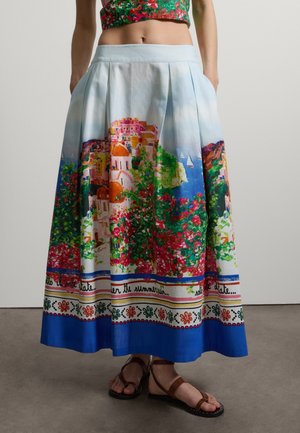 Frau trägt einen bunten Midirock mit einem Küstendorf- und Blumenmuster, kombiniert mit einem floralen Crop-Top und braunen Sandalen.