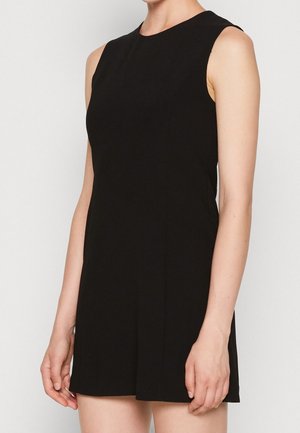 Robe noire sans manches à col montant portée par une personne debout devant un fond clair uni.